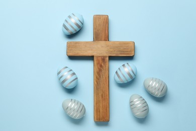 Wooden cross and painted Easter eggs on light blue background, flat lay