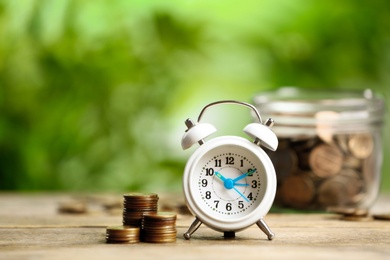 Photo of White alarm clock and stacked coins on wooden table against blurred background. Money savings