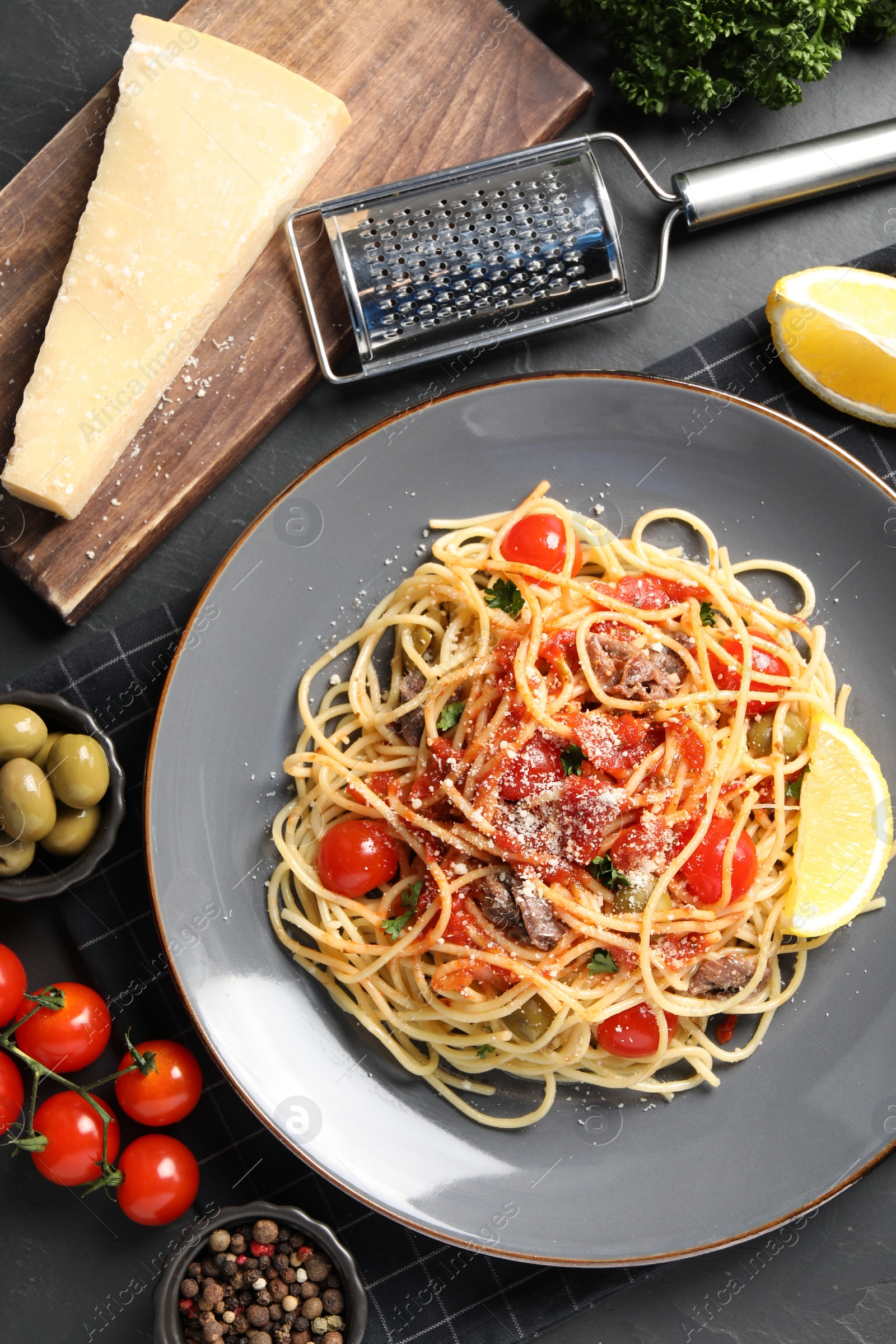 Photo of Plate of delicious pasta with anchovies, tomatoes and parmesan cheese near ingredients on black table, flat lay