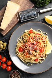 Photo of Plate of delicious pasta with anchovies, tomatoes and parmesan cheese near ingredients on black table, flat lay