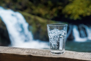 Photo of Glass of fresh water on wooden surface near waterfall. Space for text