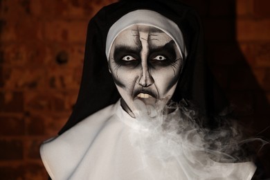 Photo of Scary devilish nun blowing smoke near brick wall. Halloween party look