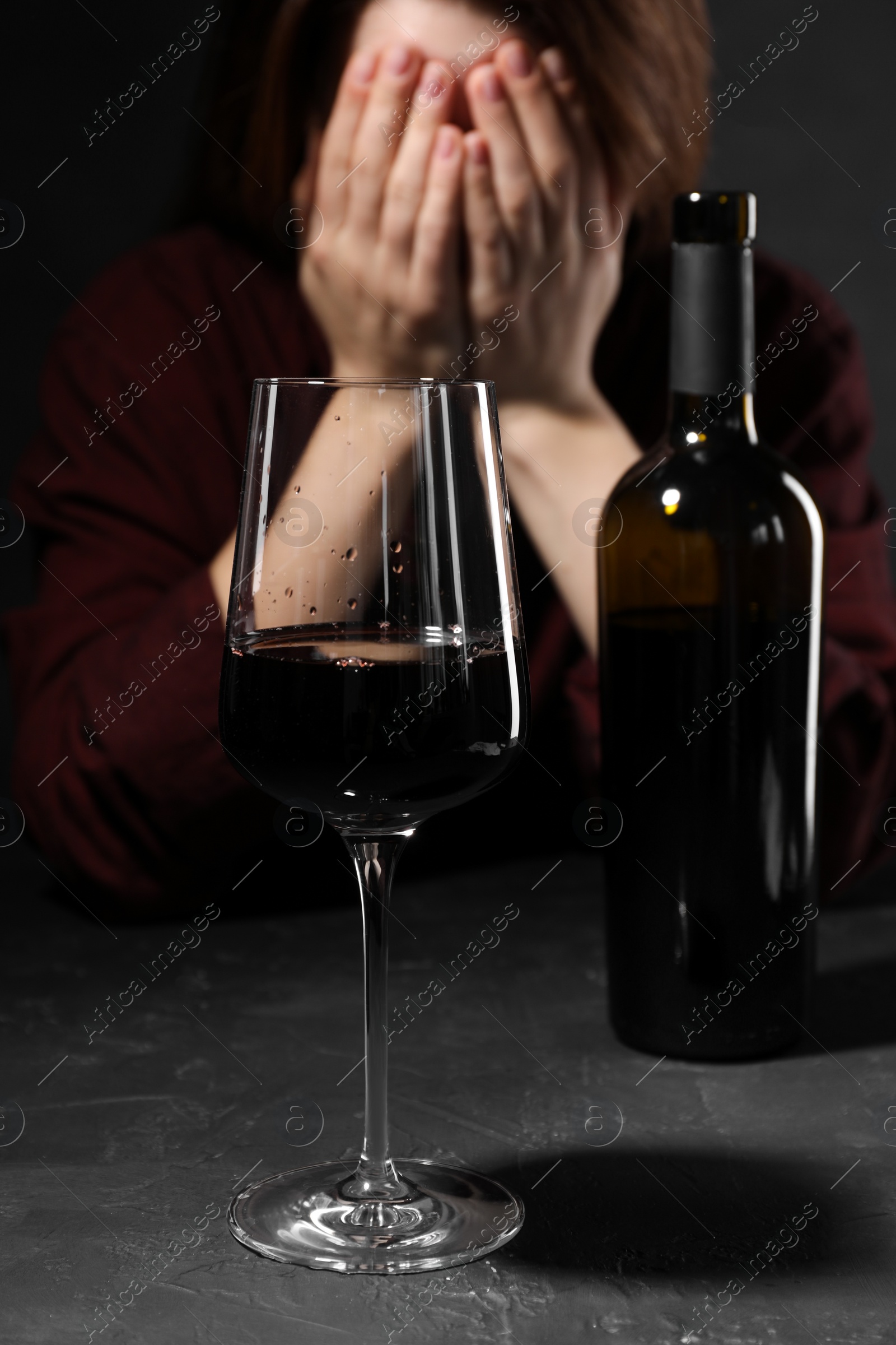 Photo of Alcohol addiction. Woman covering her face at dark textured table, focus on glass of red wine