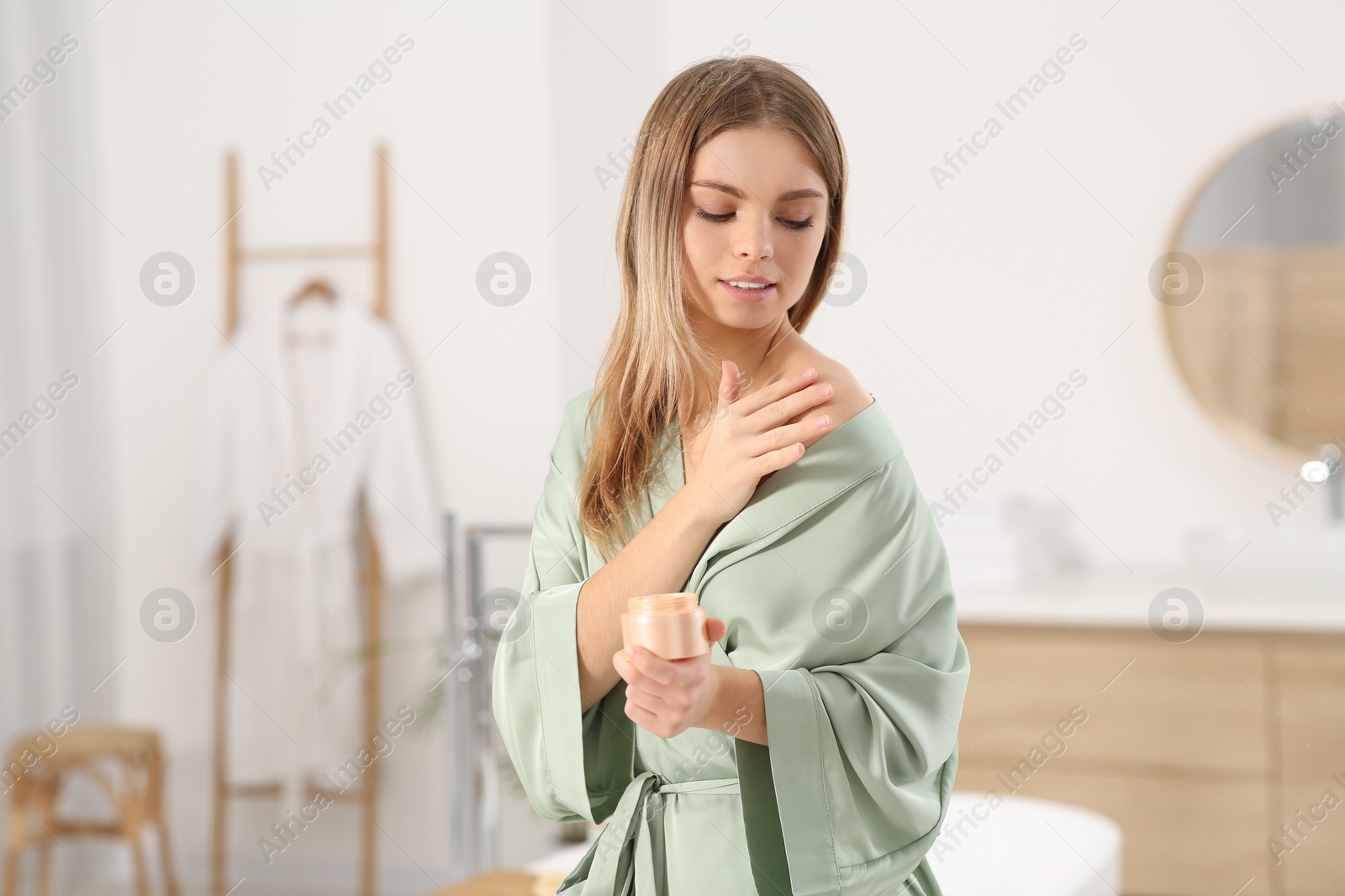 Photo of Beautiful woman with jar of cream wearing stylish robe in bathroom