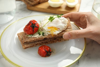 Photo of Woman eating fresh rye crispbread with poached egg, cream cheese and grilled tomato at table, closeup