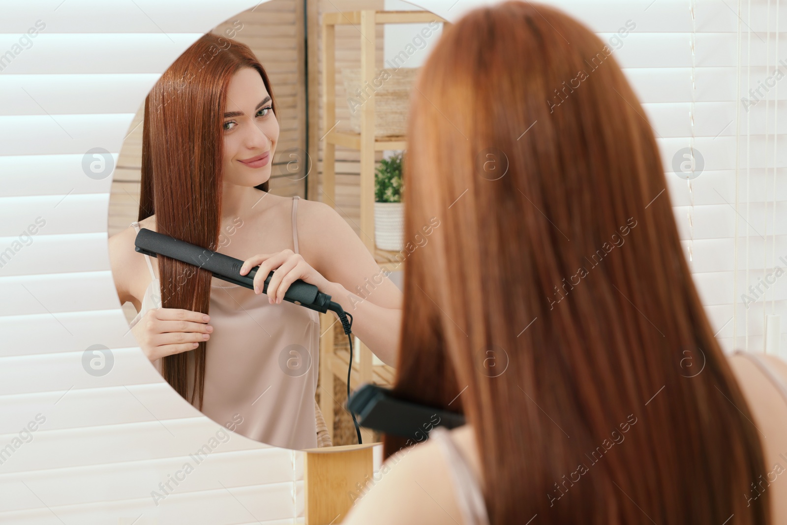 Photo of Beautiful woman using hair iron near mirror in room