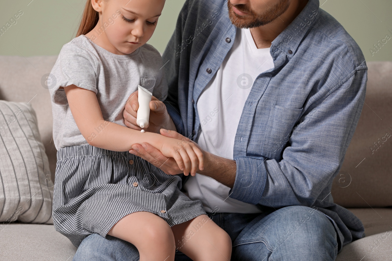 Photo of Father applying ointment onto his daughter's arm on couch, closeup