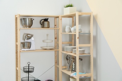 Wooden shelving units with kitchenware near color wall. Stylish room interior