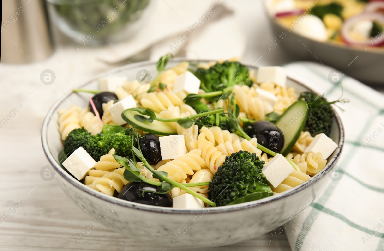 Photo of Bowl of delicious pasta with cucumber, olives, broccoli and cheese on white wooden table, closeup