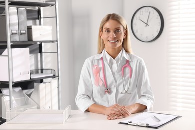 Doctor with stethoscope and pink ribbon at white desk indoors. Breast cancer awareness