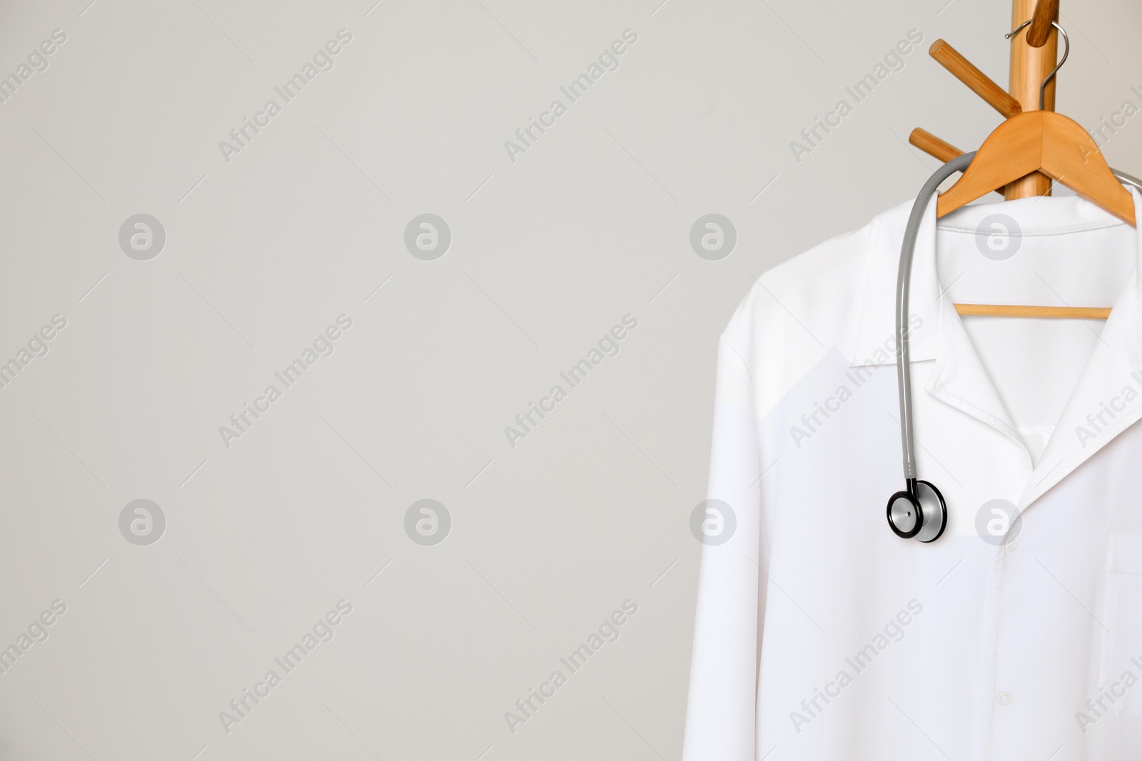 Photo of Medical uniform and stethoscope hanging on rack against light grey background. Space for text