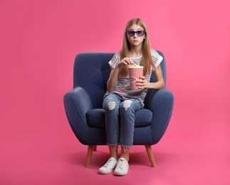 Emotional teenage girl with 3D glasses and popcorn sitting in armchair during cinema show on color background