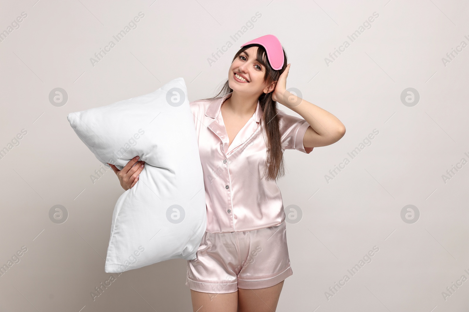 Photo of Happy woman in pyjama and sleep mask holding pillow on light grey background