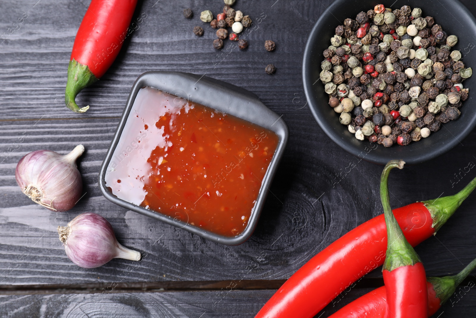 Photo of Spicy chili sauce, garlic, peppers and peppercorns on black wooden table, flat lay