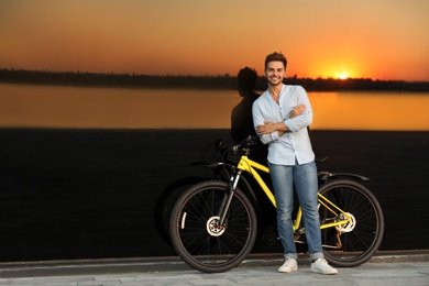 Handsome young man with bicycle near glass building at sunset. Space for text