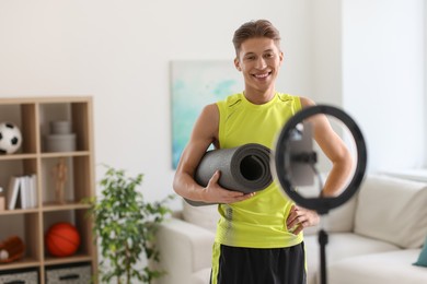 Photo of Smiling sports blogger holding yoga mat while streaming online fitness lesson at home
