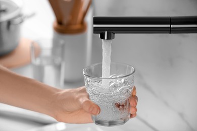 Photo of Woman filling glass with tap water from faucet in kitchen, closeup