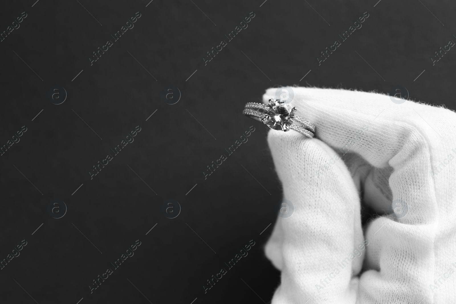 Photo of Jeweler holding diamond ring on dark grey background, closeup. Space for text