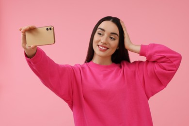 Smiling young woman taking selfie with smartphone on pink background, space for text