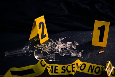 Photo of Broken bottle, yellow tape and evidence markers on black slate table. Crime scene