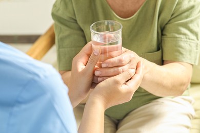 Caretaker giving glass of water to elderly woman indoors, closeup