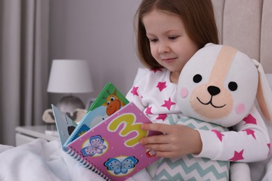Cute little girl with toy reading book in bed at home