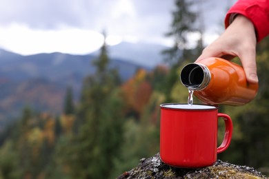 Photo of Woman pouring hot water from thermo bottle into mug in mountains, closeup. Space for text