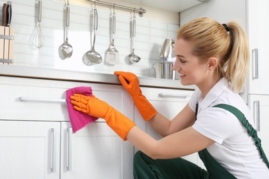 Photo of Female janitor cleaning cabinet with rag in kitchen