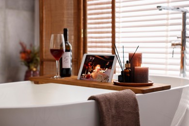 Photo of Wooden tray with tablet, wine and burning candles on bathtub in bathroom