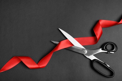 Photo of Stylish scissors and red ribbon on black background, flat lay with space for text. Ceremonial tape cutting