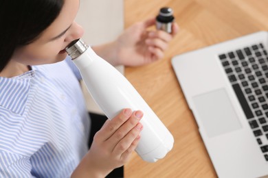 Young woman drinking from thermo bottle at workplace indoors, above view