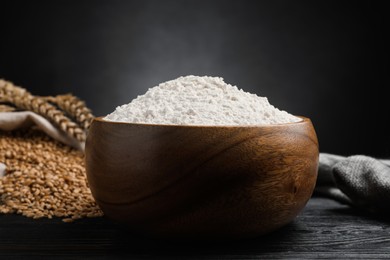 Wheat flour in bowl, spikes and grains on black wooden table, closeup