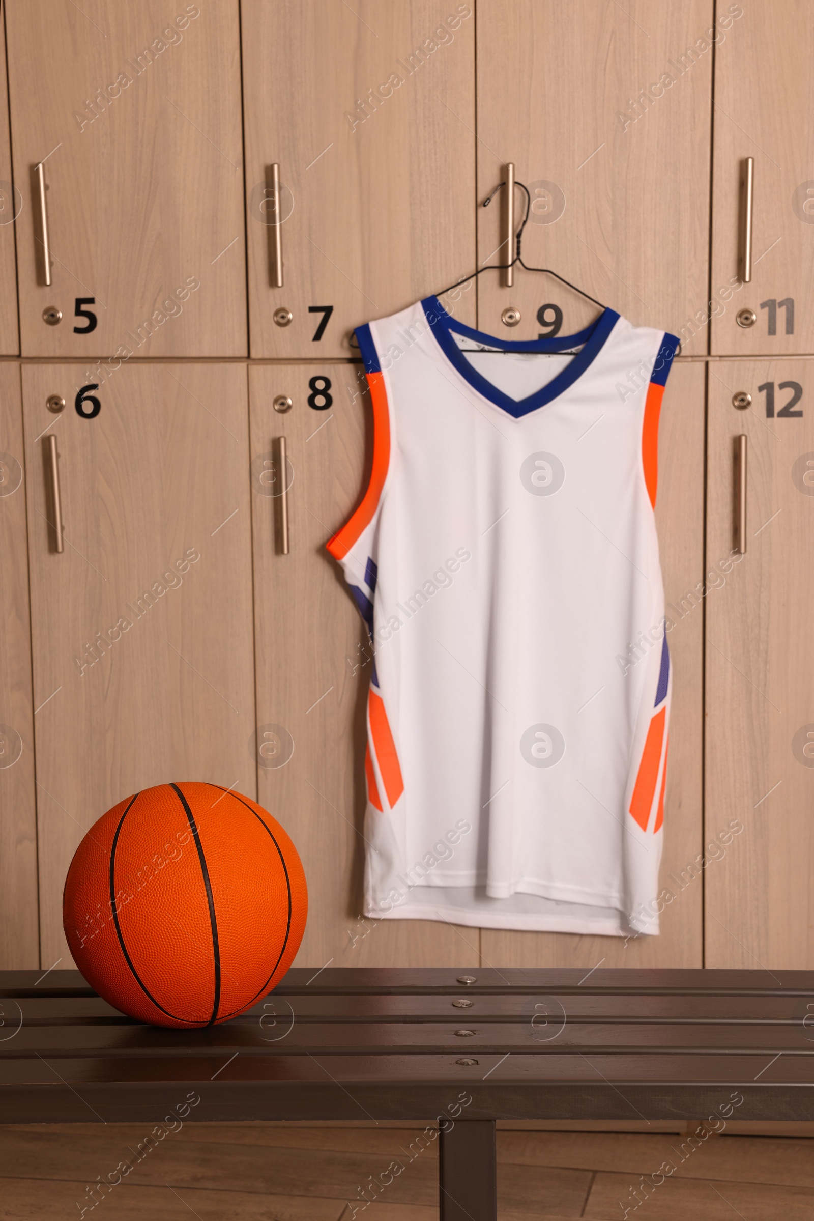 Photo of Orange basketball ball on wooden bench and hanger with uniform in locker room