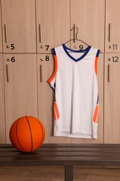 Photo of Orange basketball ball on wooden bench and hanger with uniform in locker room