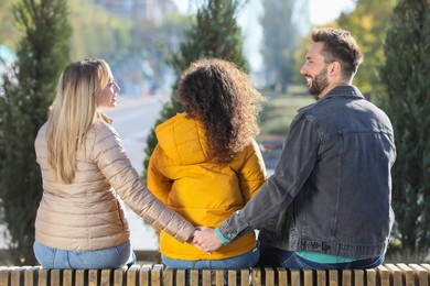 Photo of Man holding hands with another woman behind his girlfriend's back on bench in park. Love triangle