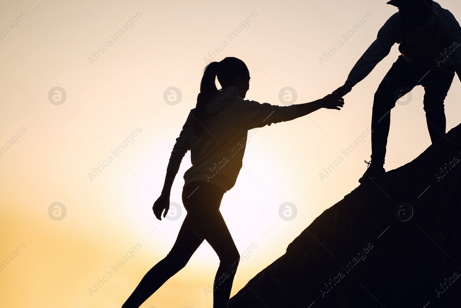 Photo of Silhouettes of man and woman helping each other to climb on hill against sunset