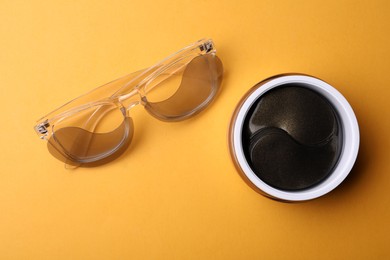 Glasses and jar with under eye patches on orange background, flat lay. Cosmetic product