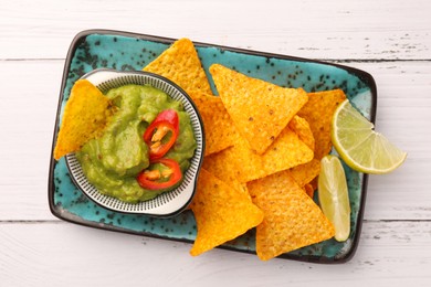 Photo of Bowl of delicious guacamole with chili pepper, nachos chips and lime on white wooden table, top view