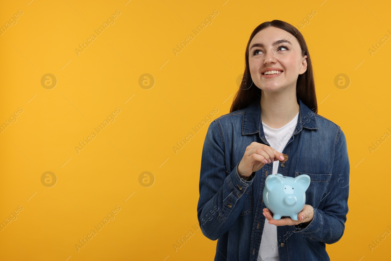 Photo of Happy woman putting coin into piggy bank on orange background, space for text