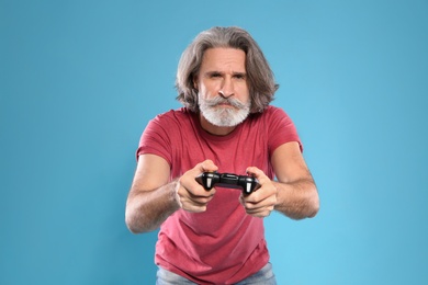 Emotional mature man playing video games with controller on color background