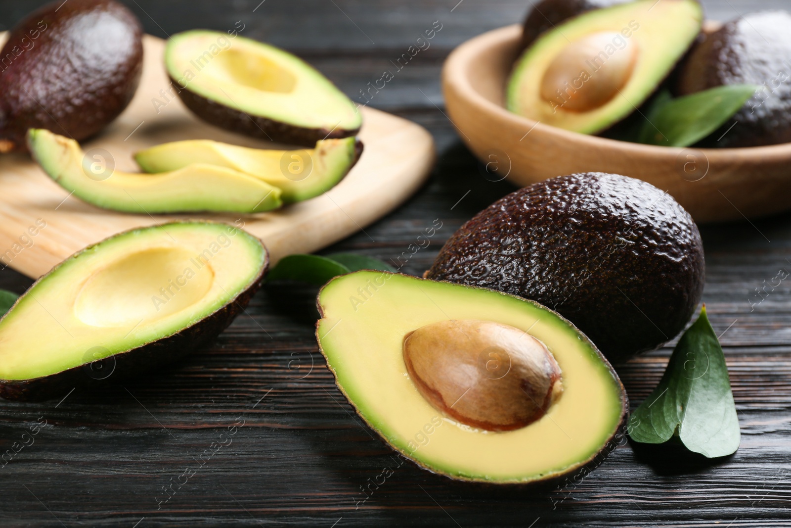 Photo of Whole and cut avocados with green leaves on dark wooden table, closeup