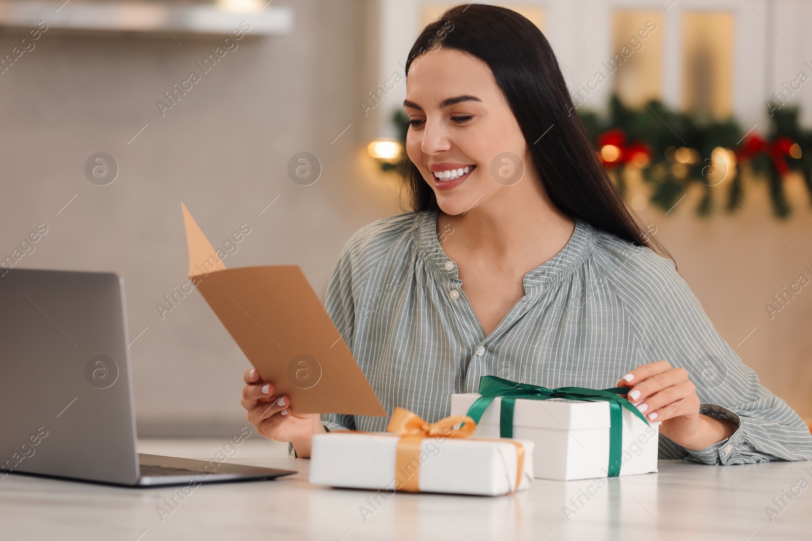 Photo of Celebrating Christmas online with exchanged by mail presents. Smiling woman reading greeting card and gifts during video call at home