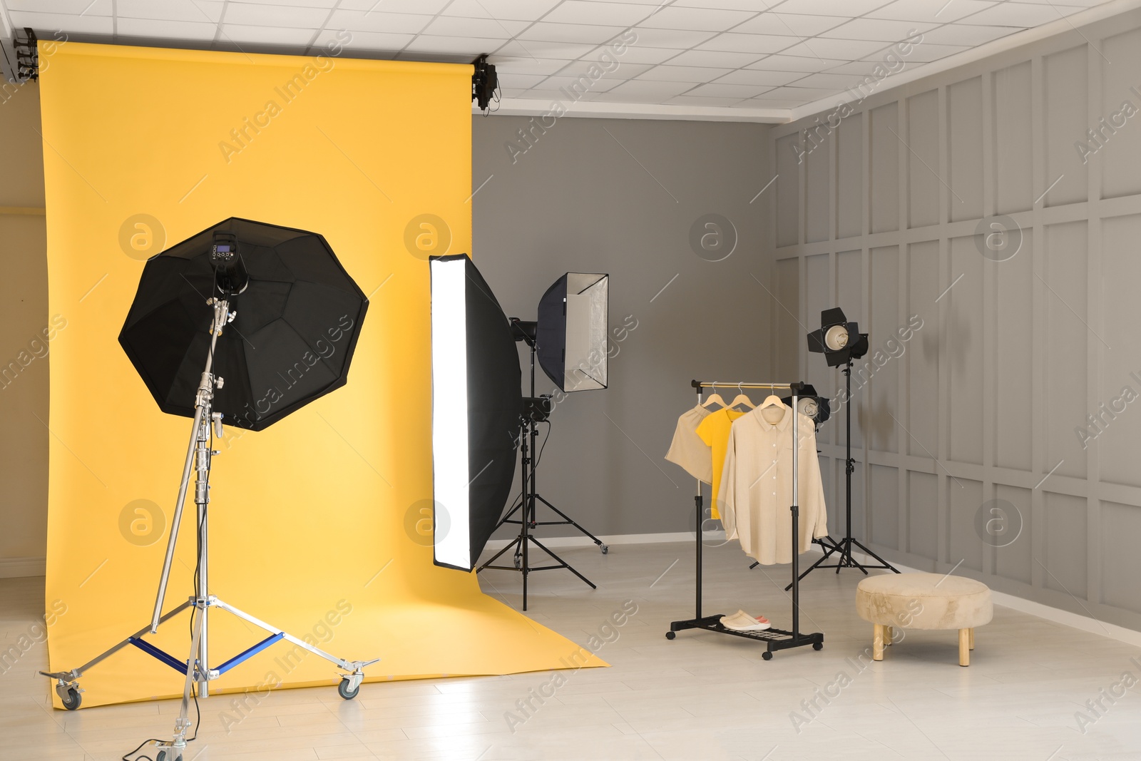 Photo of Yellow photo background, rack with clothes and professional lighting equipment in modern studio