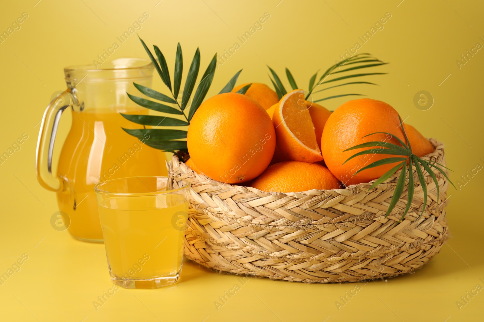 Photo of Fresh oranges in wicker basket and juice on yellow background, closeup