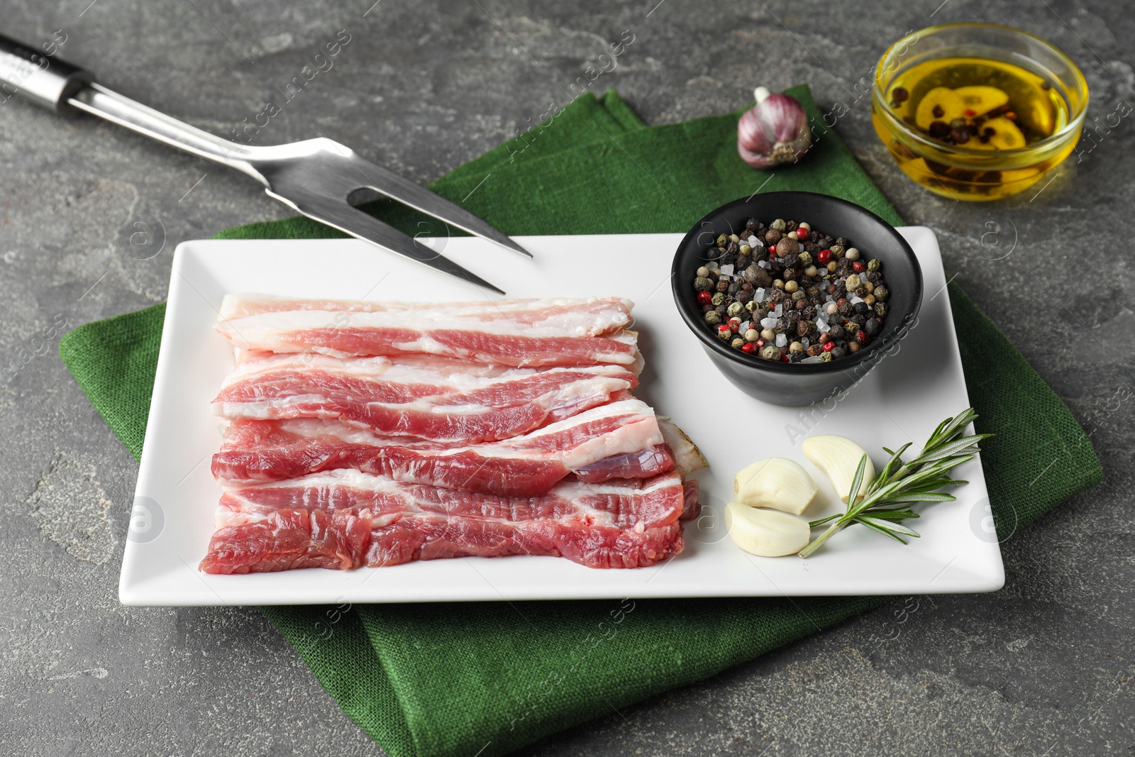 Photo of Pieces of raw pork belly, oil, peppercorns, rosemary and garlic on grey textured table