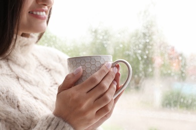 Photo of Young woman with cup of tea near window indoors on rainy day, closeup