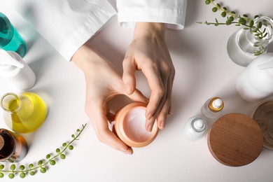 Dermatologist with jar testing cosmetic product at white table, top view