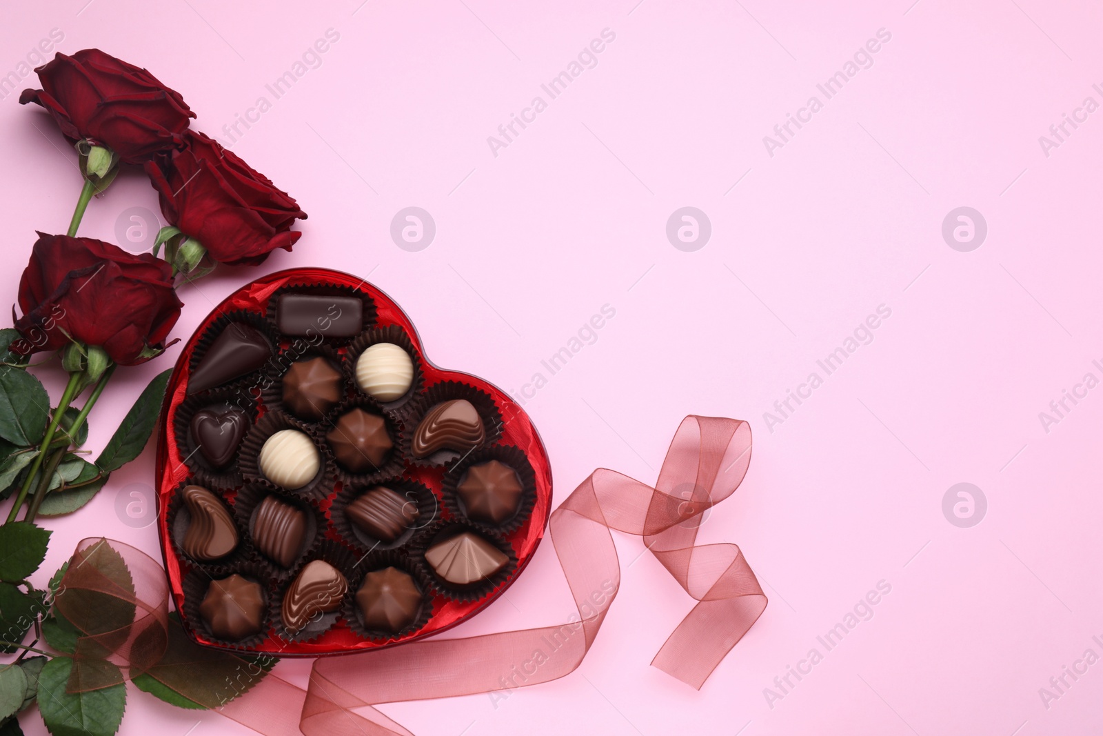 Photo of Heart shaped box with delicious chocolate candies, roses and ribbon on pink background, flat lay. Space for text