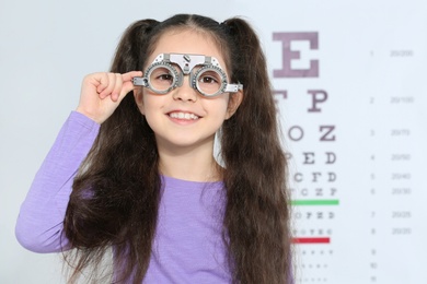 Little girl with trial frame near eye chart in hospital, space for text. Visiting children's doctor