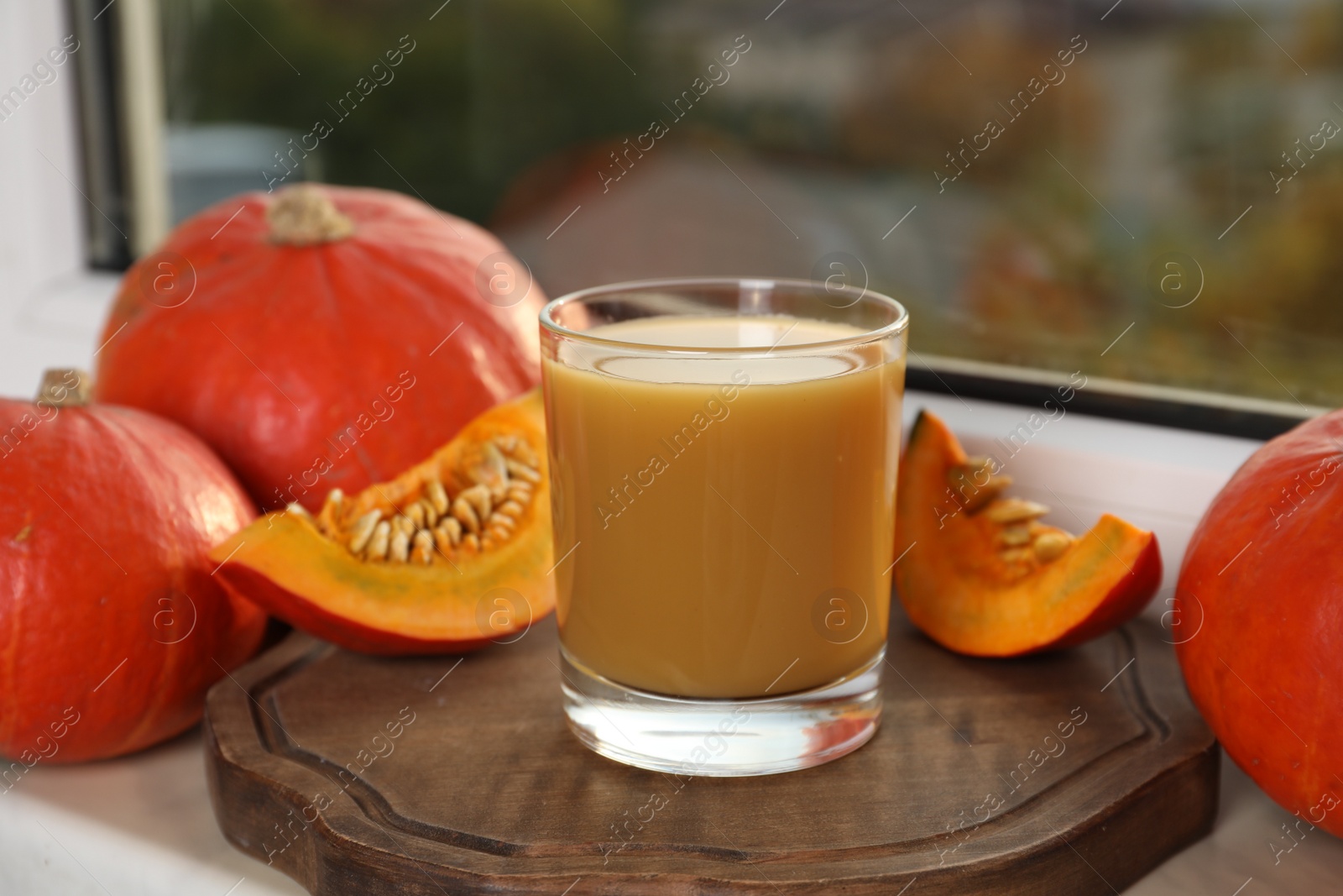 Photo of Tasty pumpkin juice in glass, whole and cut pumpkins on windowsill indoors